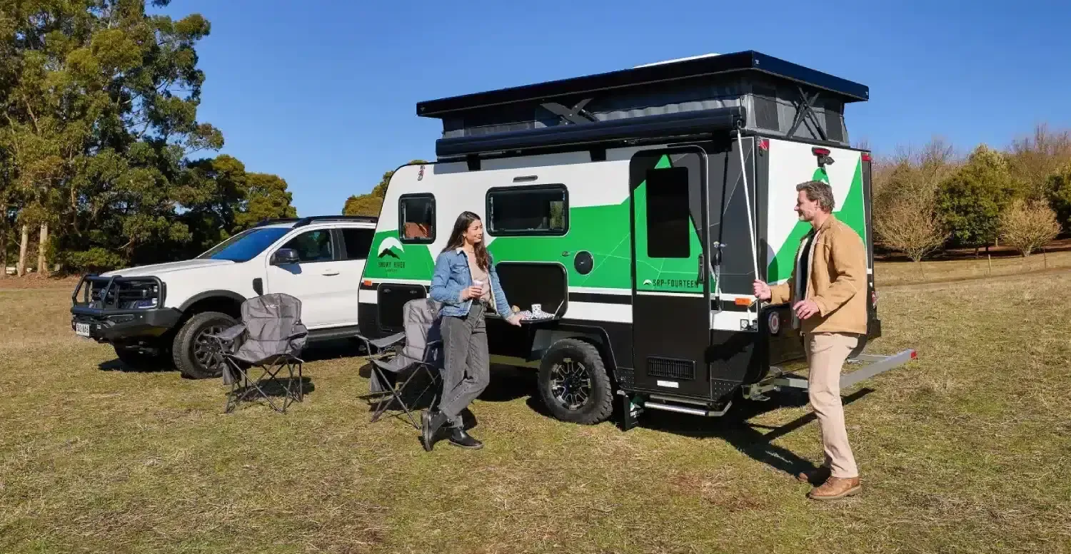 A couple beside a Snowy River pop-top camper set up in a paddock, with a 4WD parked alongside.